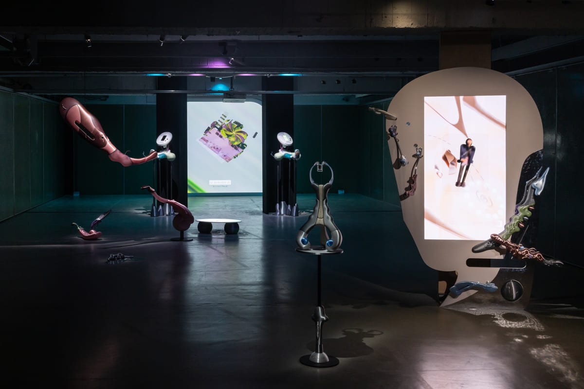 A dark exhibition room with small screens on stands, curved metallic sculptures and a large cream-coloured oval framing a bright screen.