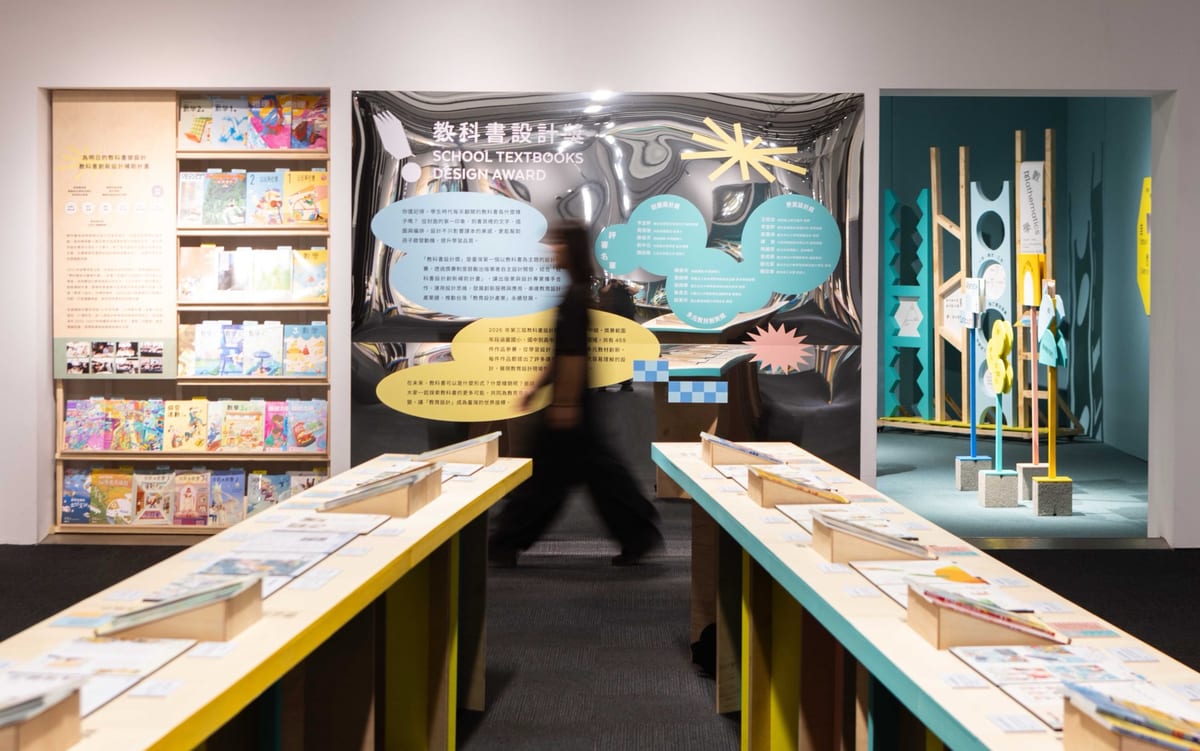 Open textbooks line two yellow tables. A blurred visitor passes between them, with bookshelves, a silver backdrop and tall coloured shapes beyond.