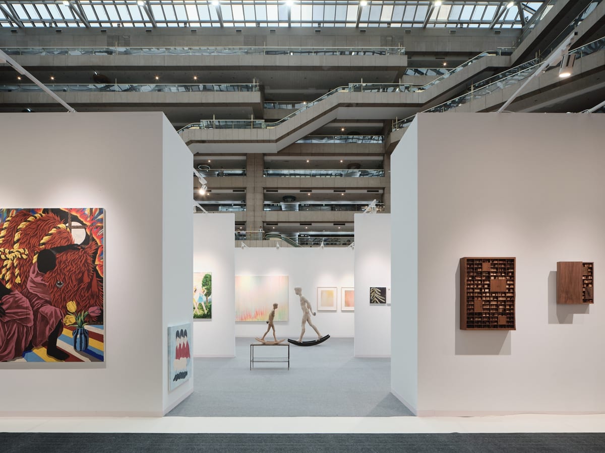 White booth walls open onto connected gallery spaces beneath the tall TWTC atrium. A vivid figure painting stands on the left, two figures balance on curved bases in the centre, and compartmented wooden works hang on the right.
