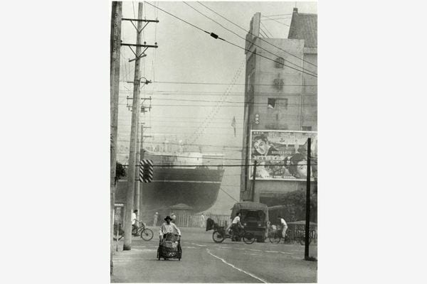 A monochrome waterfront street, with a ship behind a cinema, overhead wires and someone pushing a small cart.