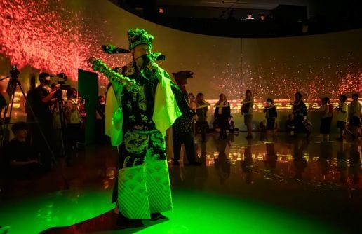 Yao Jui-Chung stands in green ritual clothing among viewers as red panoramic imagery surrounds the walls and floor.
