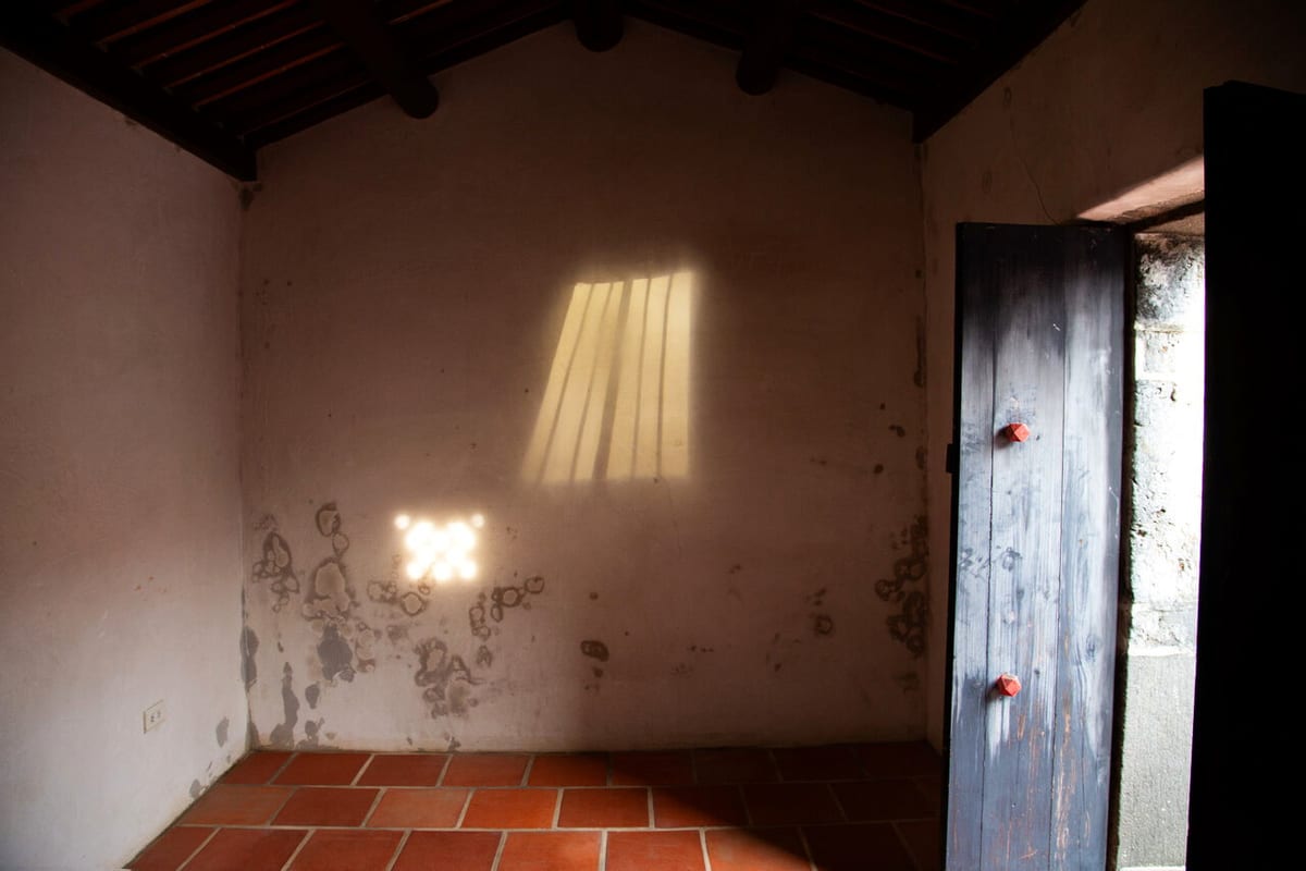 In a dark Huazhai room, sunlight makes a barred shape on the wall above a smaller pattern projected by a lamp.