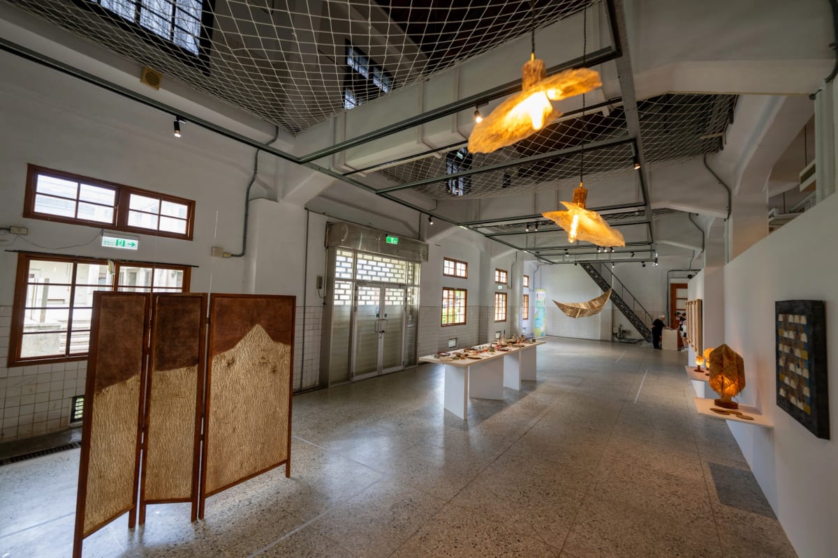 A brown fibre folding screen stands in a high-windowed gallery, with warm light passing through the bark edges of hanging and standing lamps.