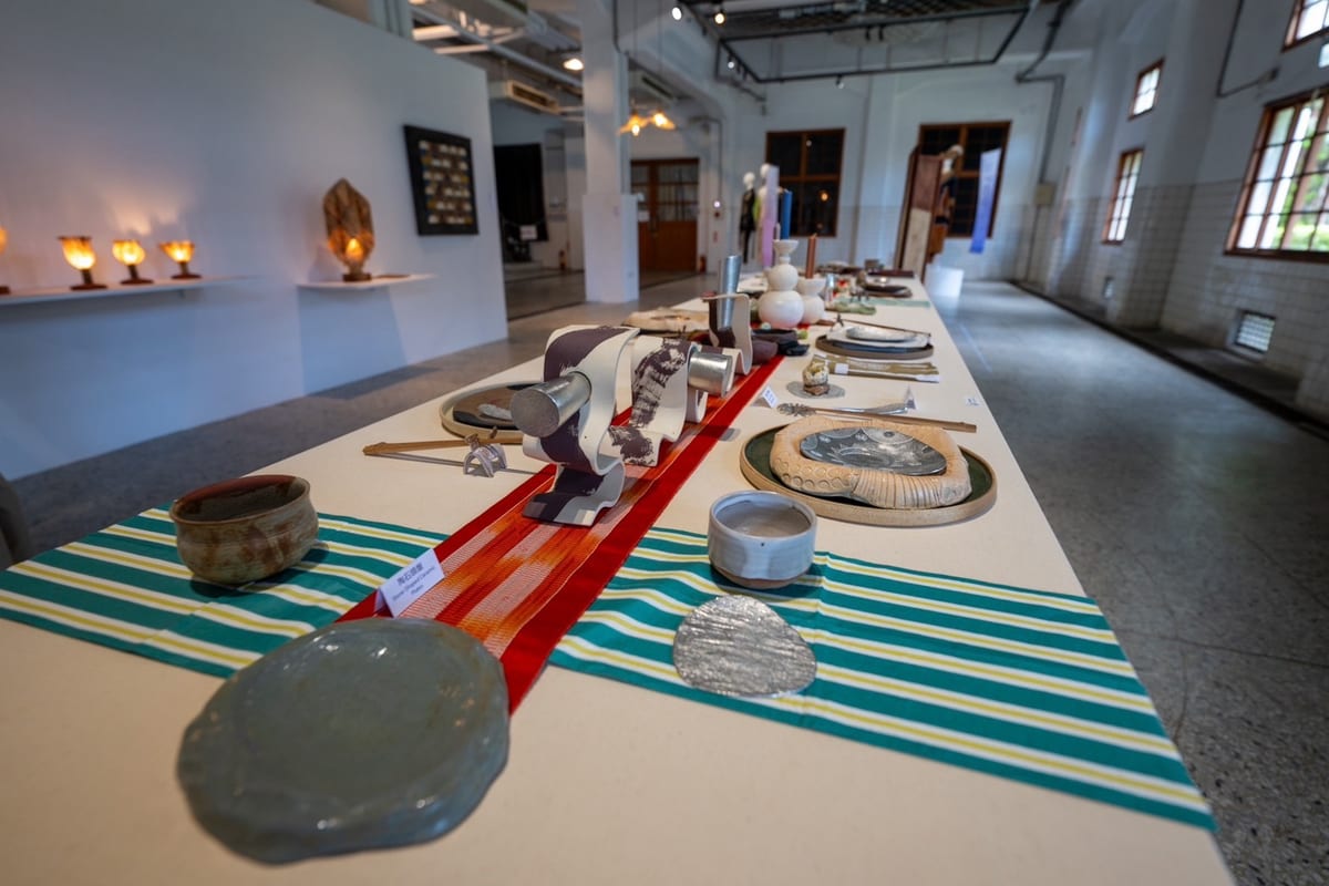 Ceramic bowls, irregular plates, tin surfaces and purple-and-white ribbon forms stretch down a table crossed by green-and-yellow cloth.