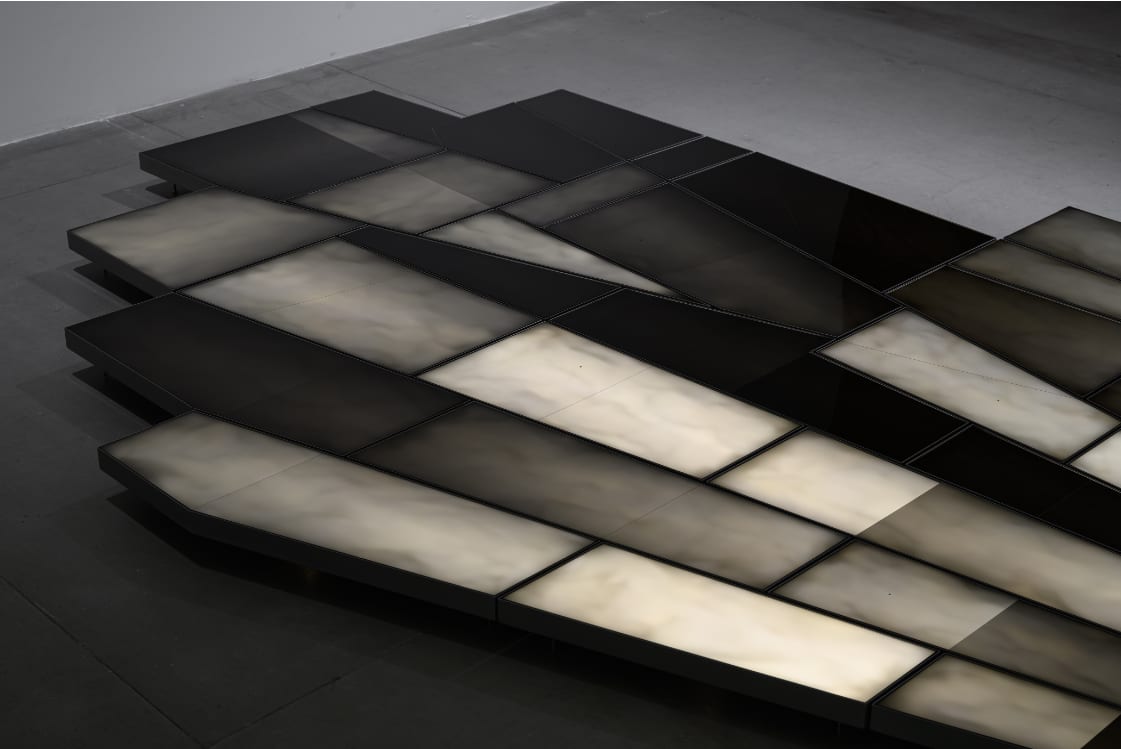 Black rectangles and pale translucent illuminated panels sit in steel frames, making a low irregular shadow-like form across a gray floor.