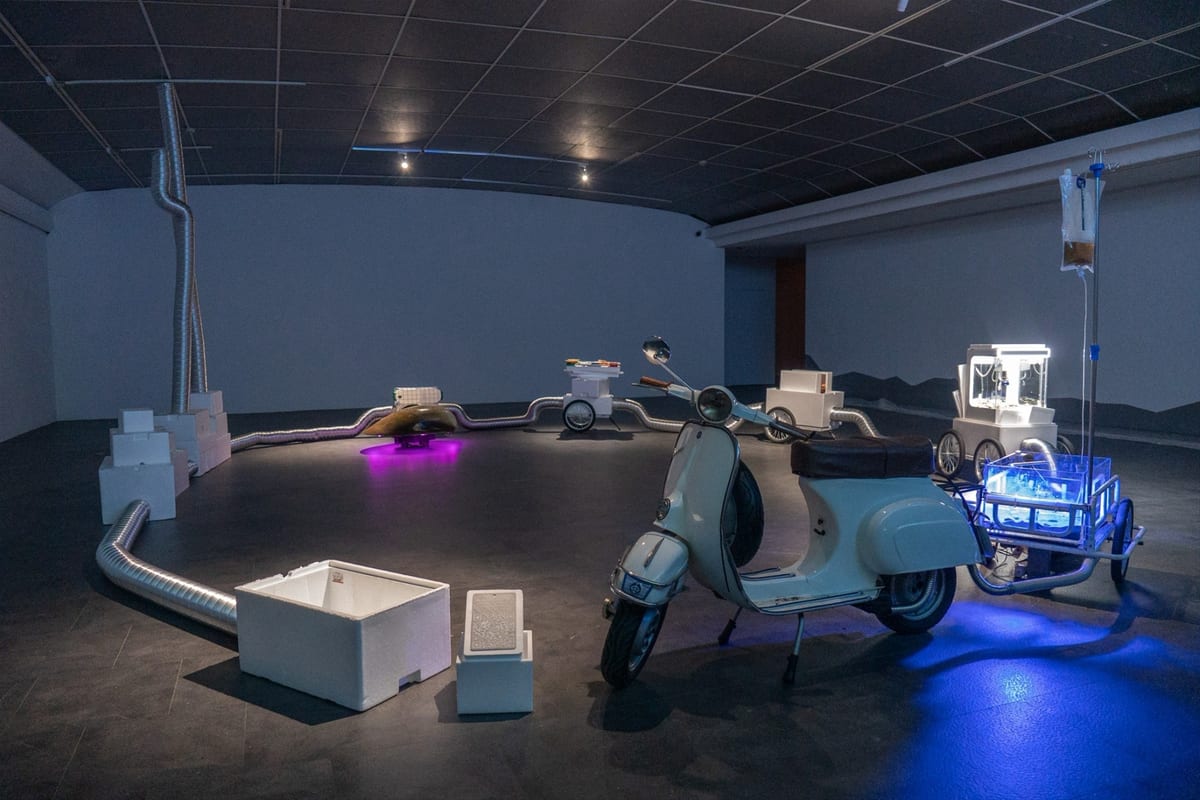 In a dim gallery, a Vespa at front right tows a trailer with a blue-lit aquarium; silver ducts loop across the floor between white foam boxes and carts, and a clam form glows in purple light at the back.