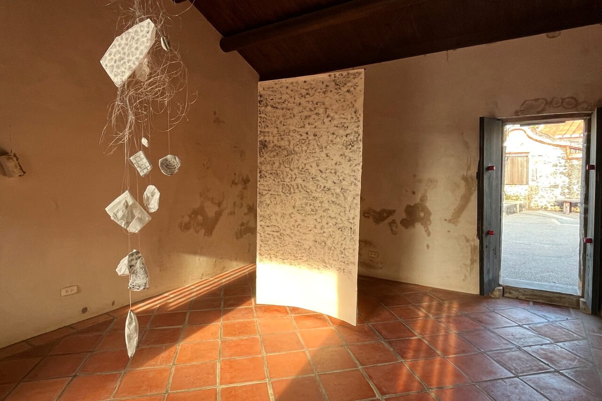 Irregular paper impressions hang beside a tall paper form as sunlight enters a Huazhai house and reaches across the red-tile floor.