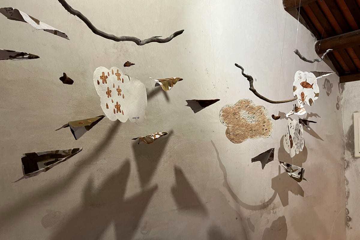 Irregular pale handmade-paper forms, folded strips, and branch-like supports hang before a wall in a Huazhai house, casting broad shadows.