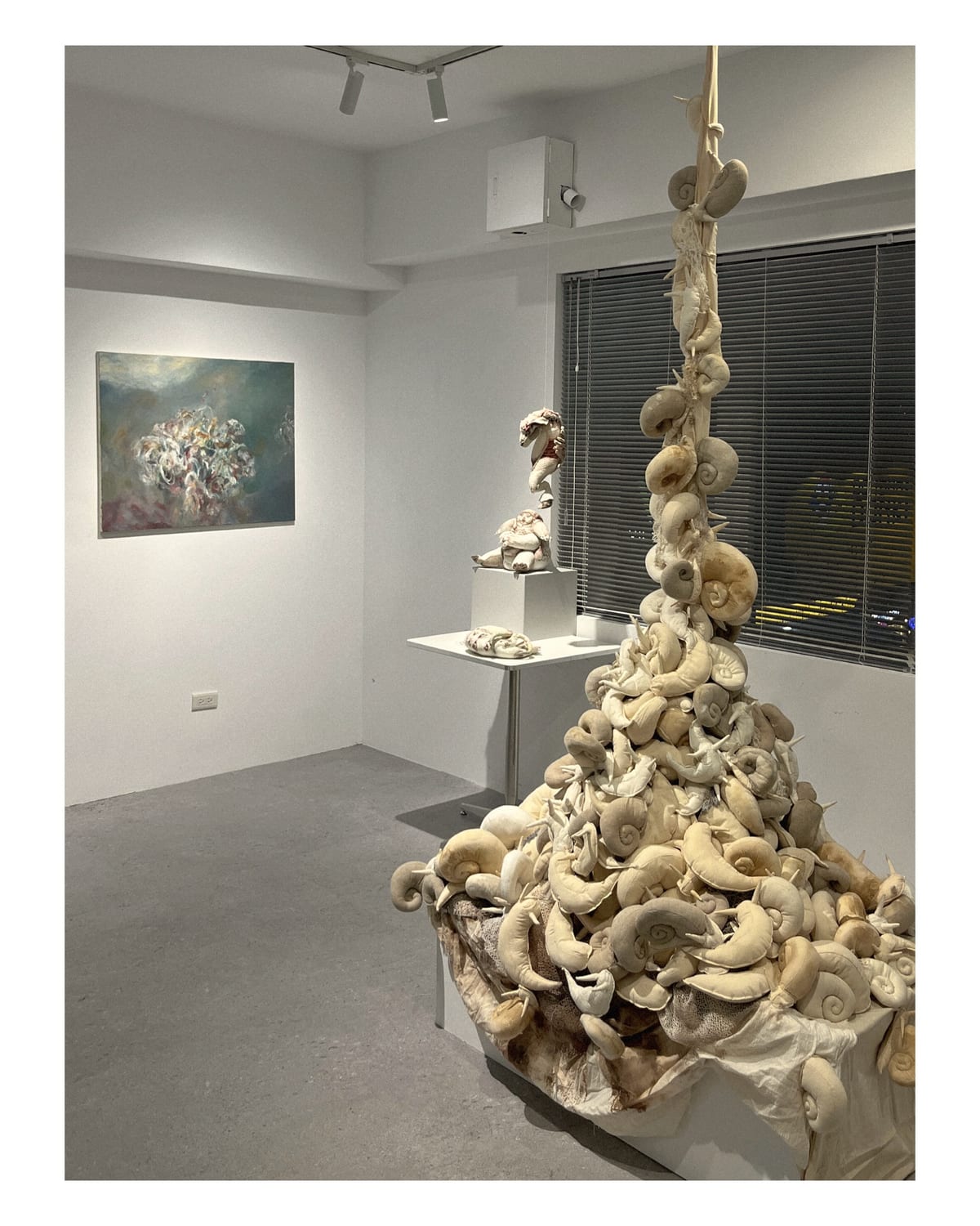 A tall hanging accumulation of pale snail-like forms fills the foreground; a painting and smaller fiber works stand beyond it.