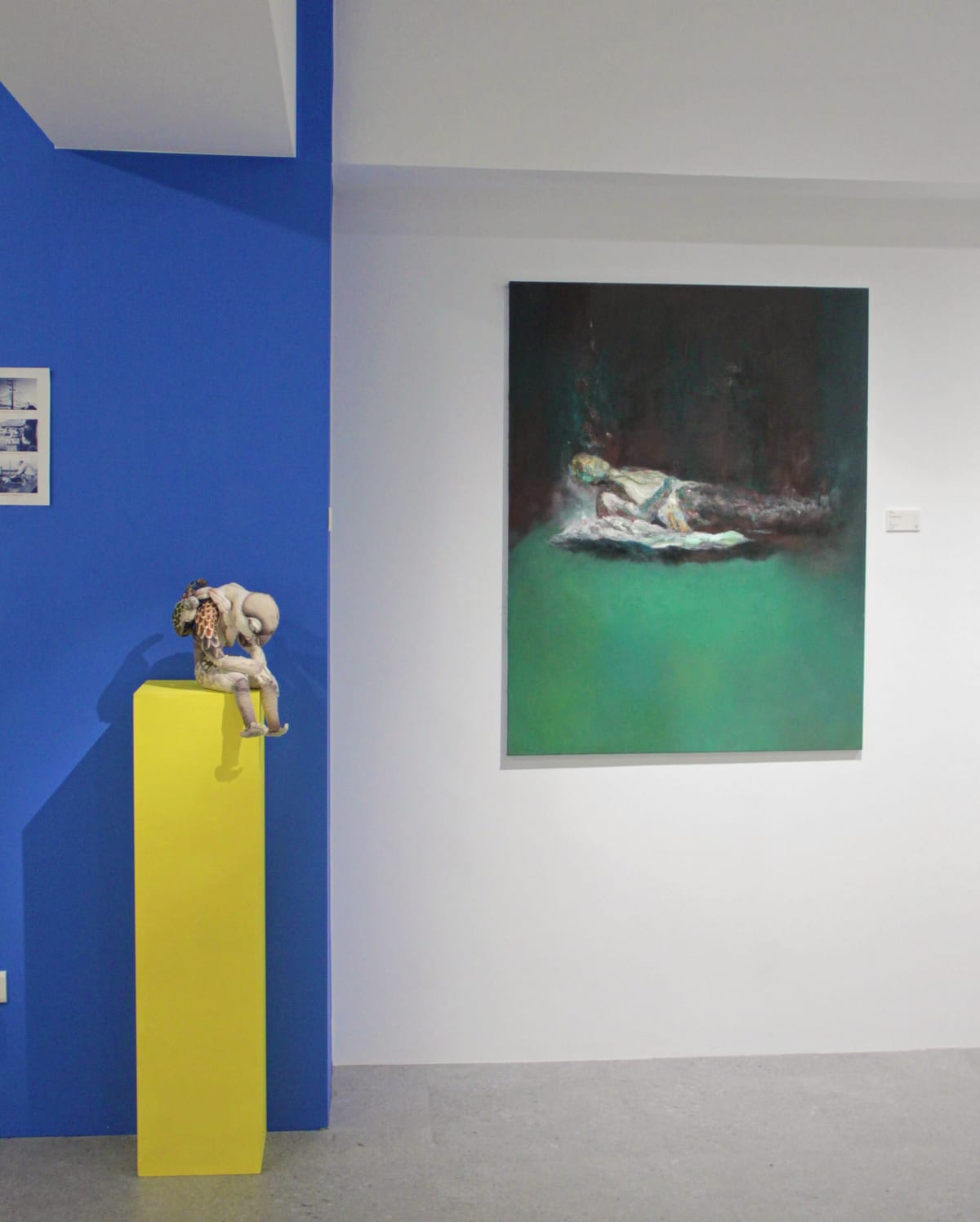 A reclining figure on a green ground hangs on a blue wall beside a small pale fiber work on a yellow plinth.