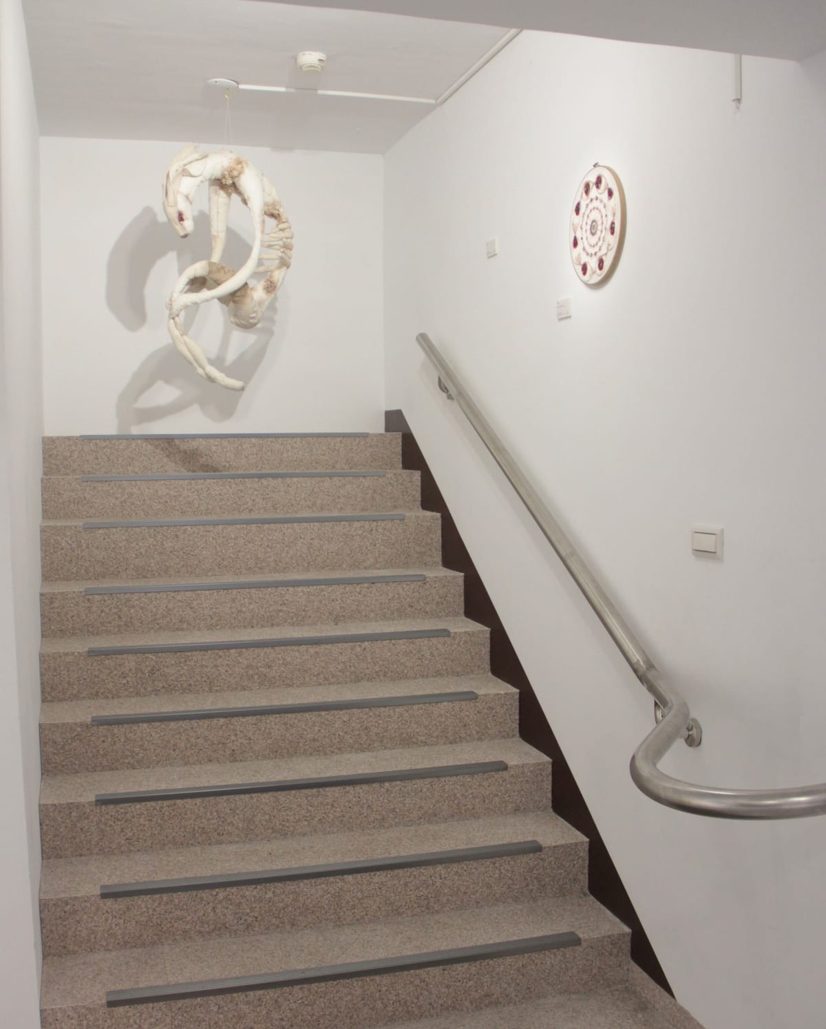 A pale fiber body bends into an unclosed loop above a stair landing, with a smaller circular fiber work on the opposite wall.