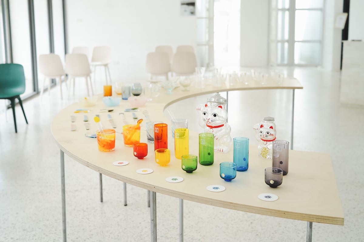 A sunlit curved wooden table holds coloured glasses, clear lucky-cat containers and small glass spoons.