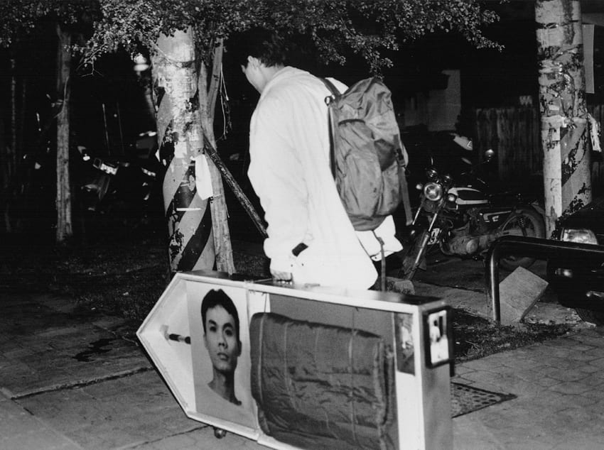 In a black-and-white photograph, Gao Jun-honn leans forward with a pack on his back, pulling an arrow-shaped wheeled box on a rope.