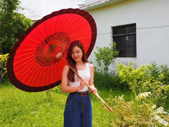 In a Meinong garden, a woman holds a large red umbrella with a black border and intricate cut-paper pattern around its centre.