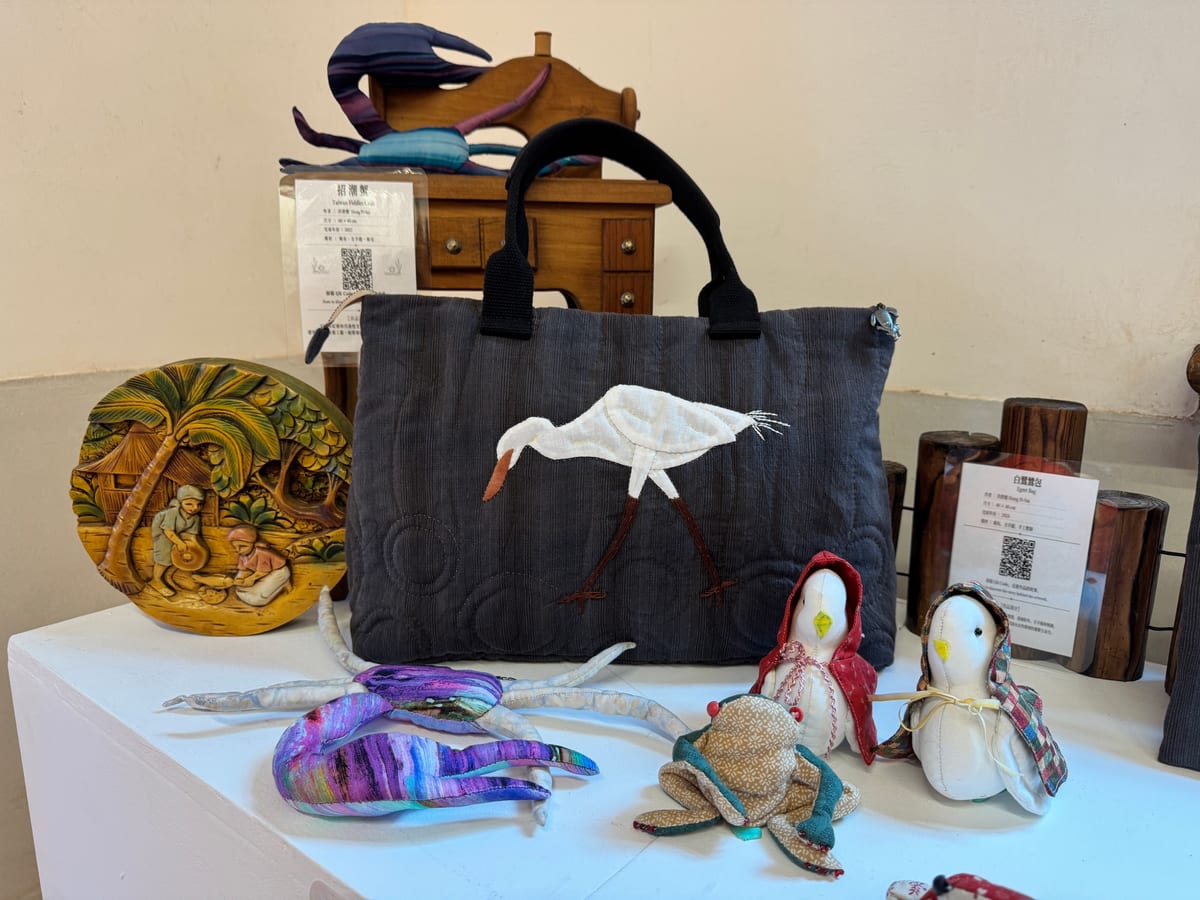 A white egret sewn onto a dark tote bag, surrounded by purple-blue striped fabric fiddler crabs and small textile figures.