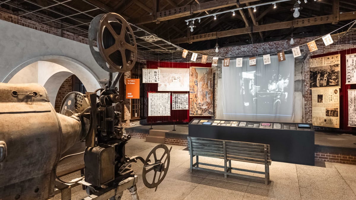 A large film projector beside a wooden bench facing a black-and-white projection, with red curtains and exposed roof beams around the reconstructed theatre.