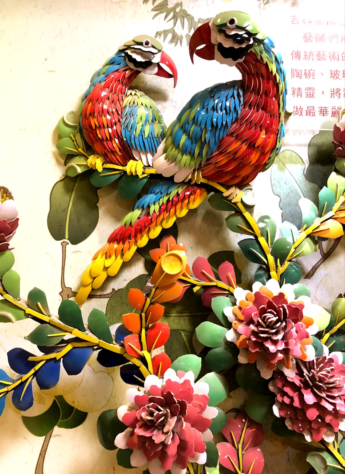 Two parrots with overlapping red, blue, yellow and green ceramic feathers above layered pink flowers and green leaves.
