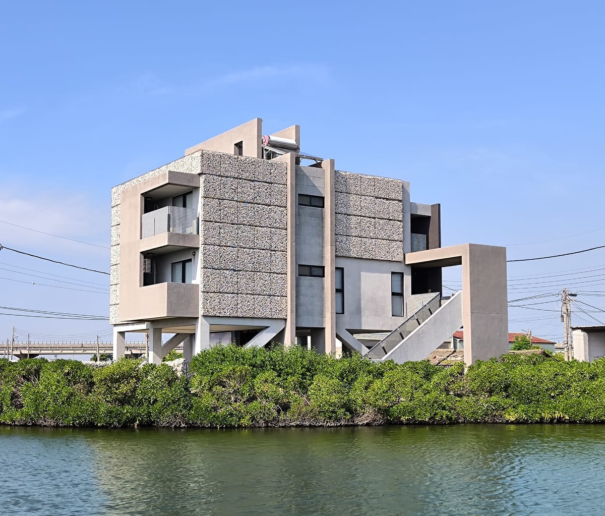 Pale oyster-shell panels on a raised house beside water, with an open lower level and external stairs.