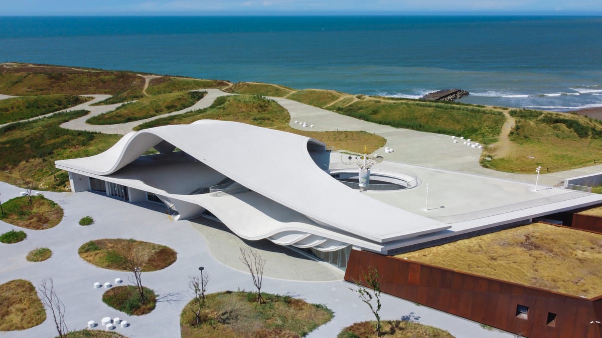 A white curving roof wraps a courtyard and broad terrace on coastal grassland, with the sea behind it.