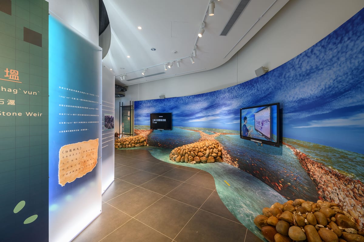 Rounded stones form low walls beneath a curved photograph of sea and stone weirs, with screens set into the display.