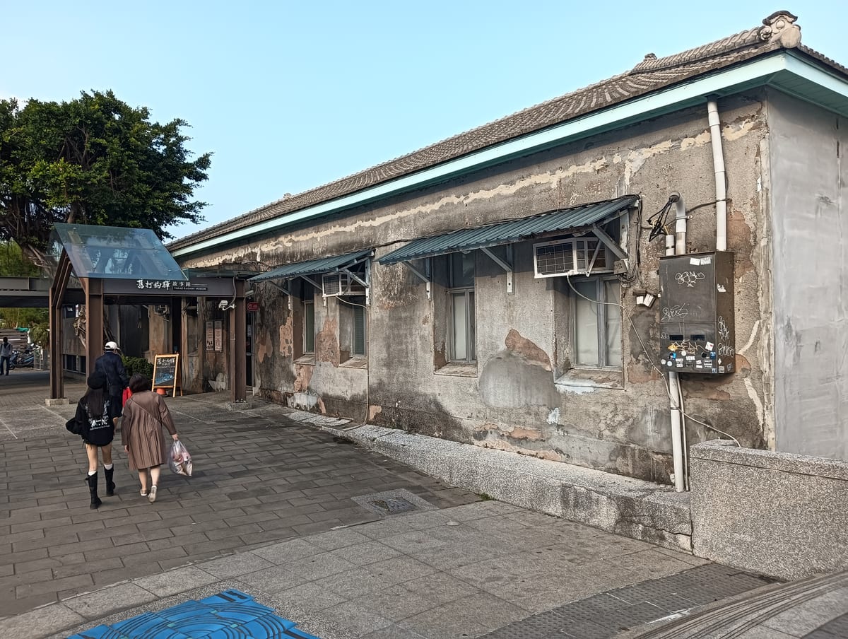 A long patched grey station wall, mint-green eaves, tiled roof and window awnings, with two visitors at the entrance.