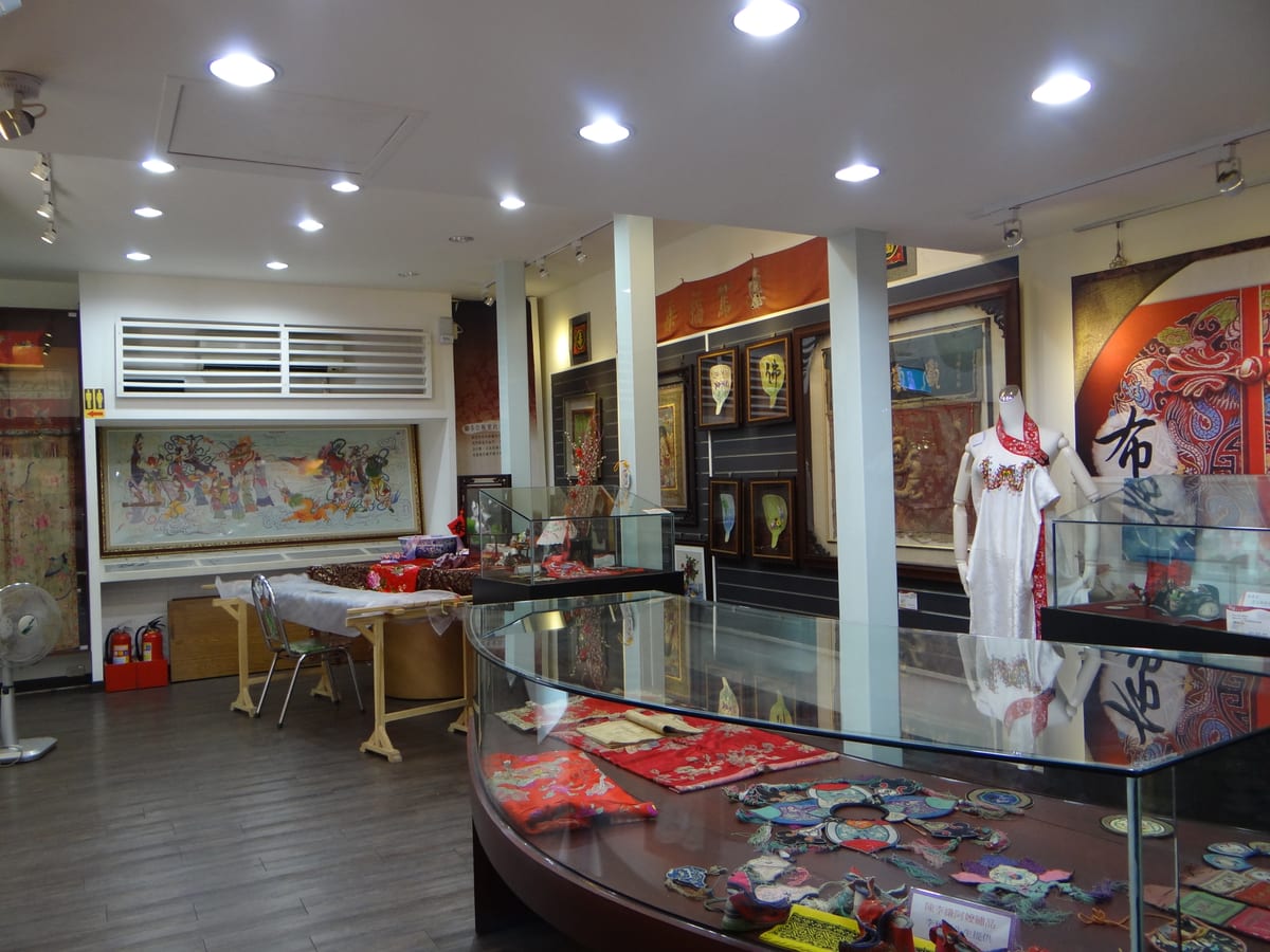A curved glass case holds small embroidered collars and garments; a mannequin in white and framed textiles fill the room behind it.