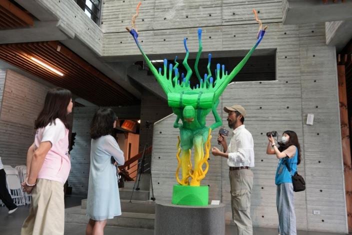 A towering green humanoid sculpture with blue-tipped limbs carries another body overhead; visitors stand beside it in the tall gallery.