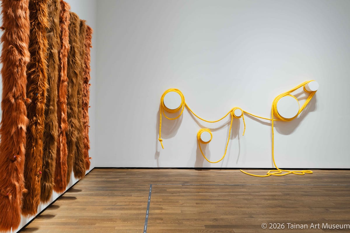 A yellow form crosses circular wall mounts, drops and gathers loosely on the floor beside a large dense brown hanging mass.