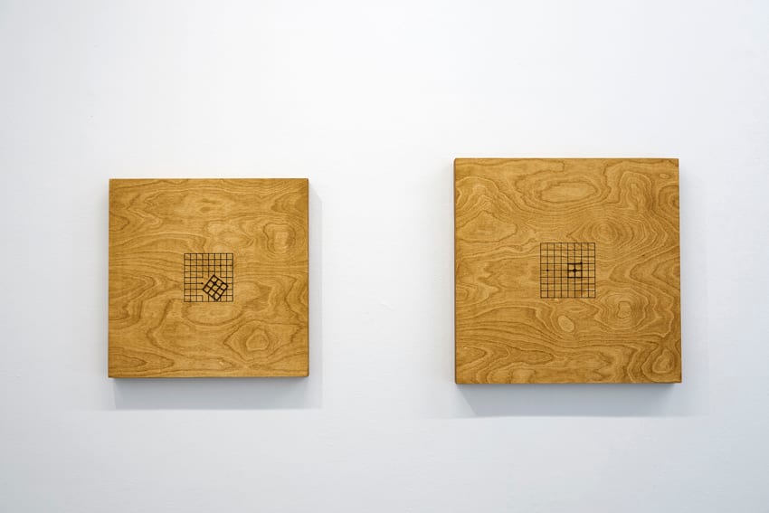 Two square stained-wood objects hang on a wall, each holding a small grid and clock movement near its centre.