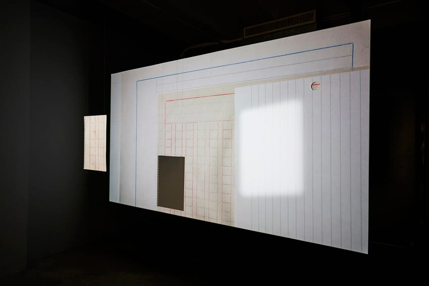 A ruled-paper field is projected inside a dark room, with lines, pale planes and an architectural shape appearing across it.