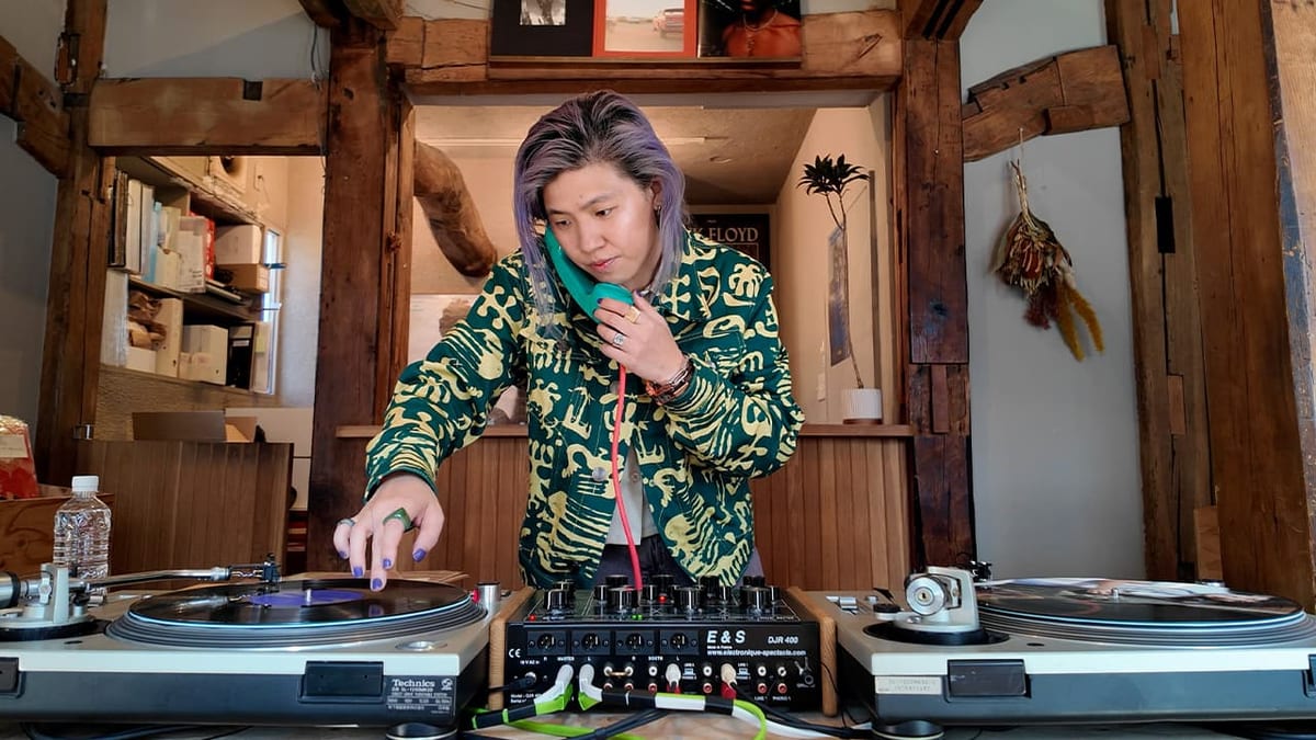 Vicki Huang stands between two turntables, one hand reaching toward the record on her left, a telephone-style headphone held to one ear.