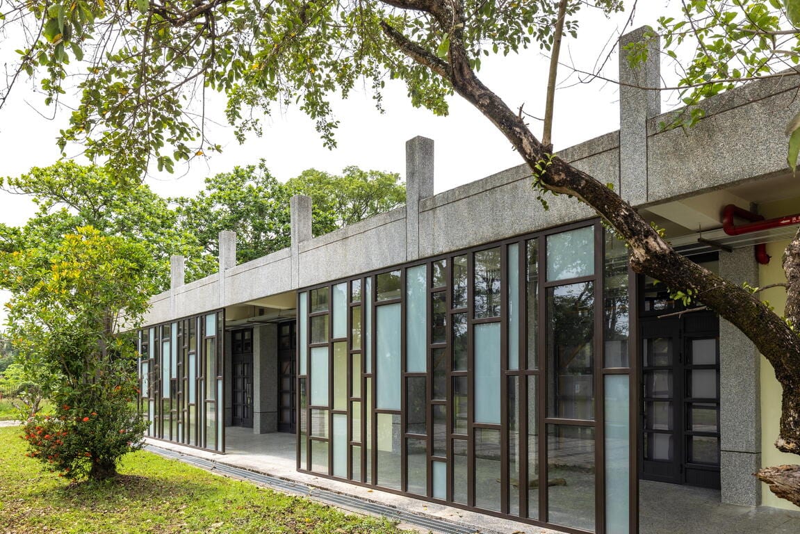 A low grey school building stands beside grass and leafy trees, with tall glass panels along its corridor.