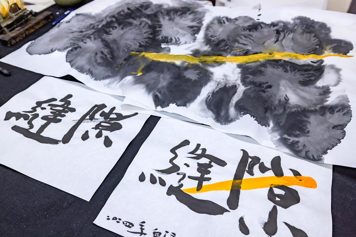 Grey-black ink spreads across white paper beneath a long yellow stroke; sheets in front carry black calligraphy.