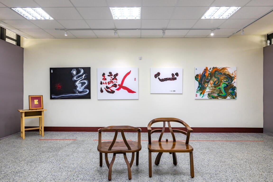 Two wooden chairs face four square calligraphy works on a white wall, with black-and-white, red-and-black, and green-and-orange strokes.