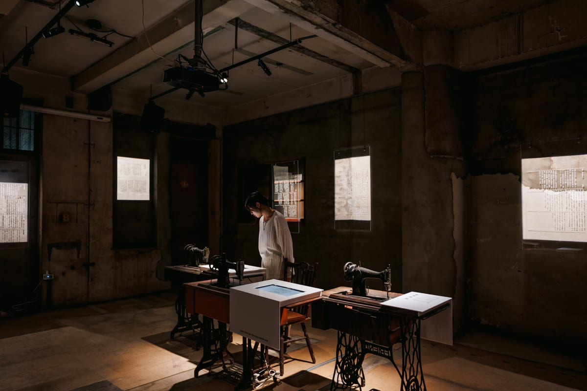A warmly lit old room: antique sewing machines on wooden tables, transparent text panels hanging above, worn walls behind.