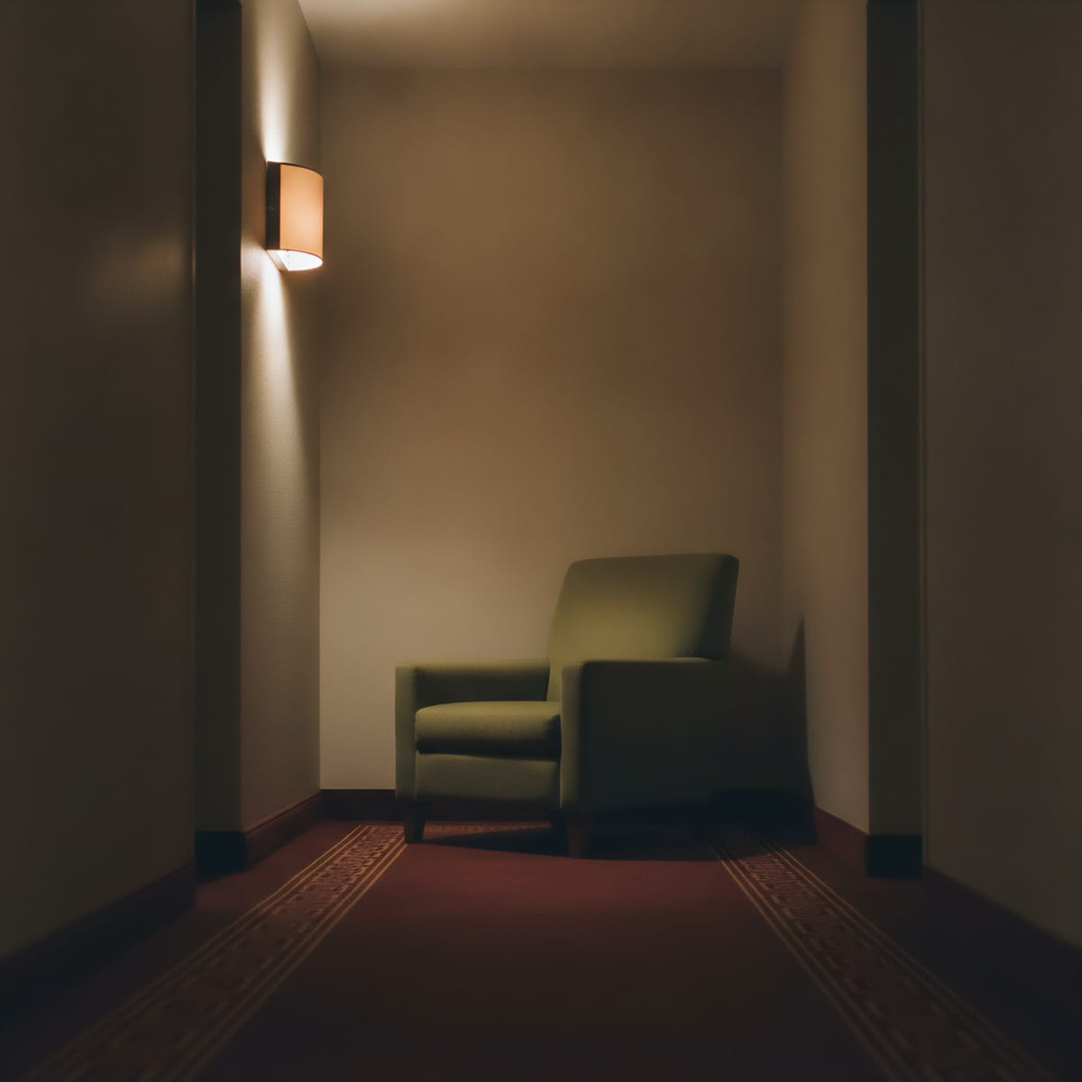 An olive-green armchair stands in a warm-brown alcove beneath a wall sconce. The light spreads up and down the wall; a patterned maroon carpet covers the floor.