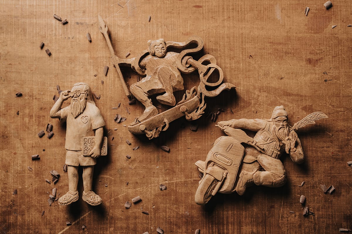 Three woodcarvings on a chip-covered work surface: Yue Lao with a tablet at left, Nezha on a dragon-shaped skateboard above, and Guan Yu on a scooter at right.
