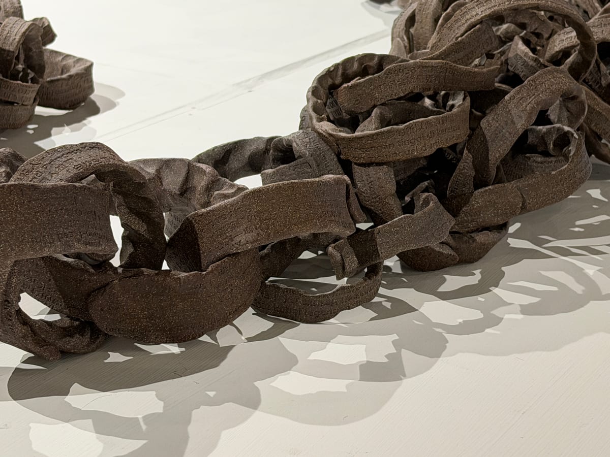Rough brown ceramic chain links, their surfaces stamped with small text, loop together in heaps on a white floor, casting shadows.