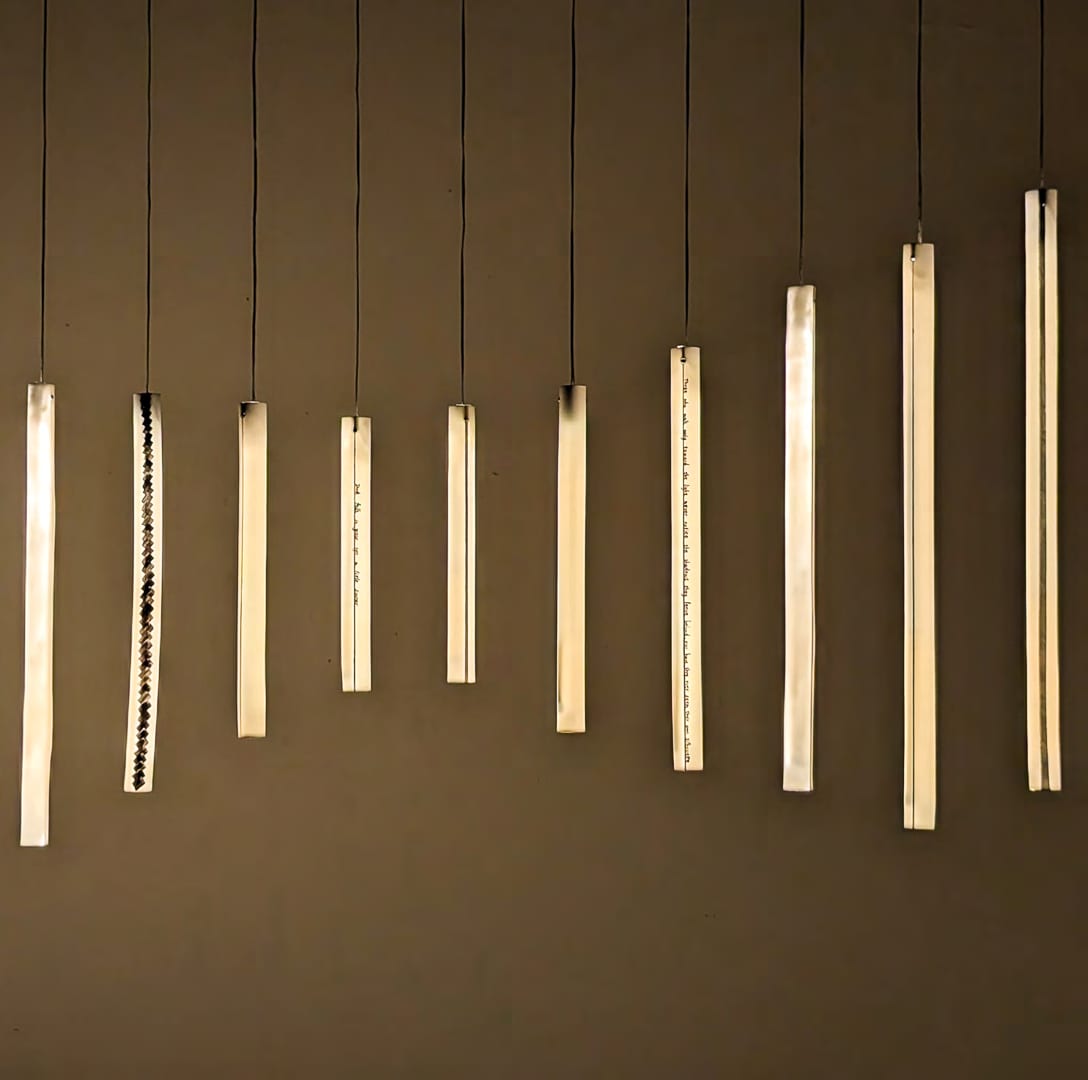 Warmly lit ceramic tubes hang at different heights against a brown wall; narrow dark glaze marks run along several surfaces.