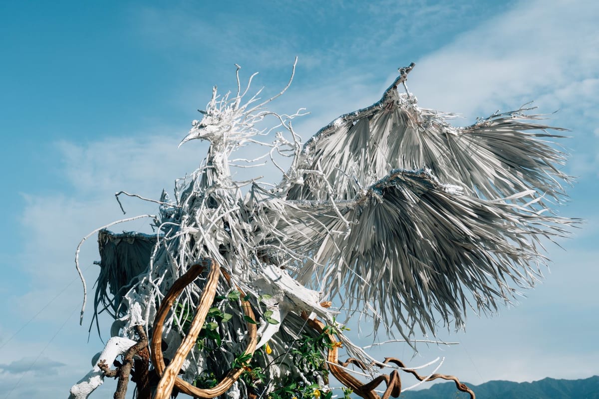 A plant phoenix stands on a concrete terrace, white branches extending from its head and wings spreading beneath a blue sky.