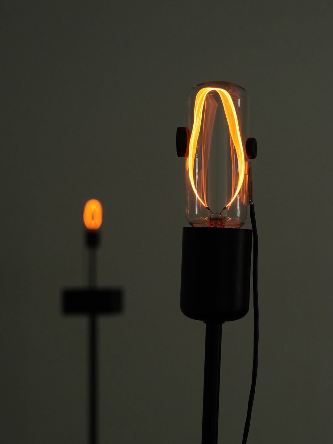 Orange filaments loop inside an upright glass tube in a dim room; a second bulb glows out of focus behind it.