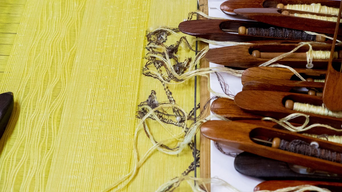 On a loom, yellow cloth woven with a floral pattern fills the left side; a row of wooden shuttles lies to the right, with pale silk threads running from them toward the cloth.