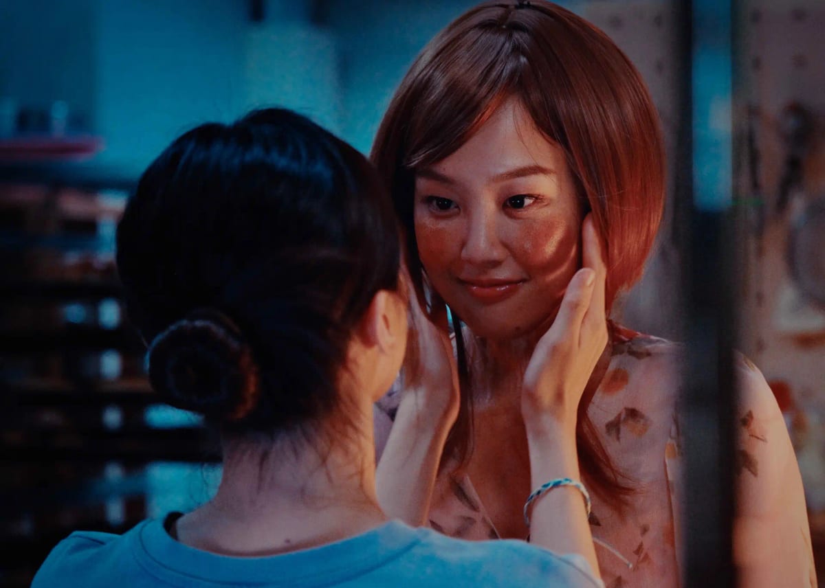 Against blue light, a dark-haired woman faces away from the camera and holds a short-haired woman's face in both hands. Brown patches and fine creases mark the face and neck in warm light.