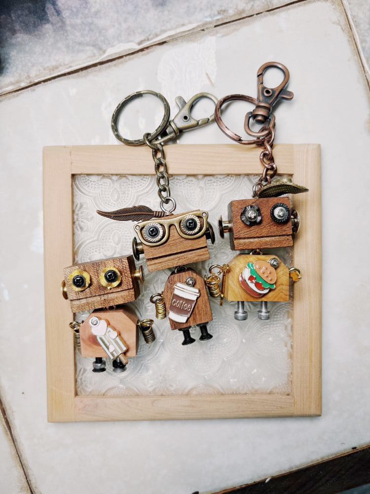 Three wooden robot keyrings with screw eyes, spring arms and small drink or hamburger decorations rest on a framed lace background.