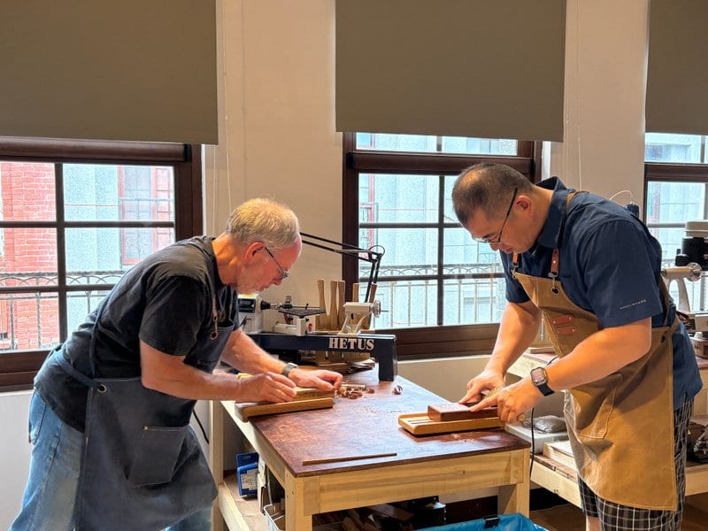 Two people in aprons lean over a wooden workbench by tall windows, using hand planes on strips of wood. Wood shavings lie on the bench.