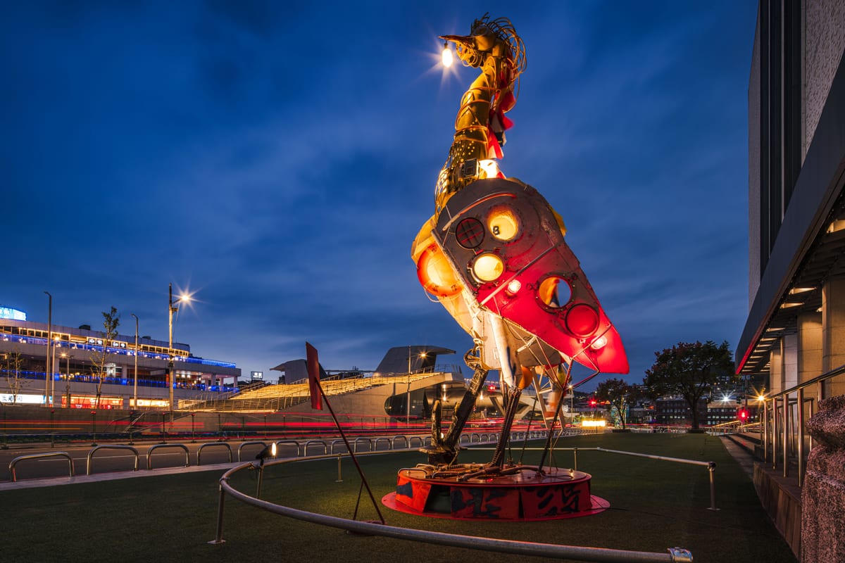 A tall metal egret glows beside Keelung Art Museum at dusk. Round ship windows form its body, chains form its legs, and a light hangs from its beak.