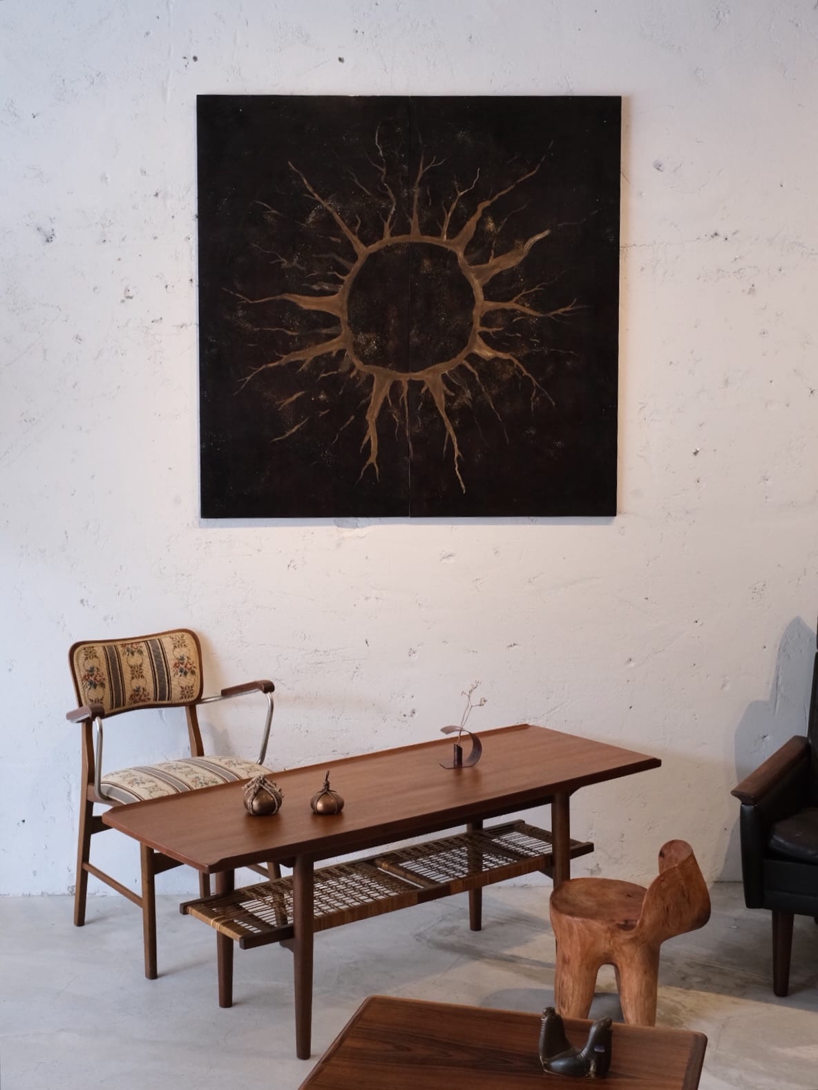 Brown branching forms radiate around a hollow circle in a dark square work; a wooden table, patterned chair and small objects sit below against a white wall.