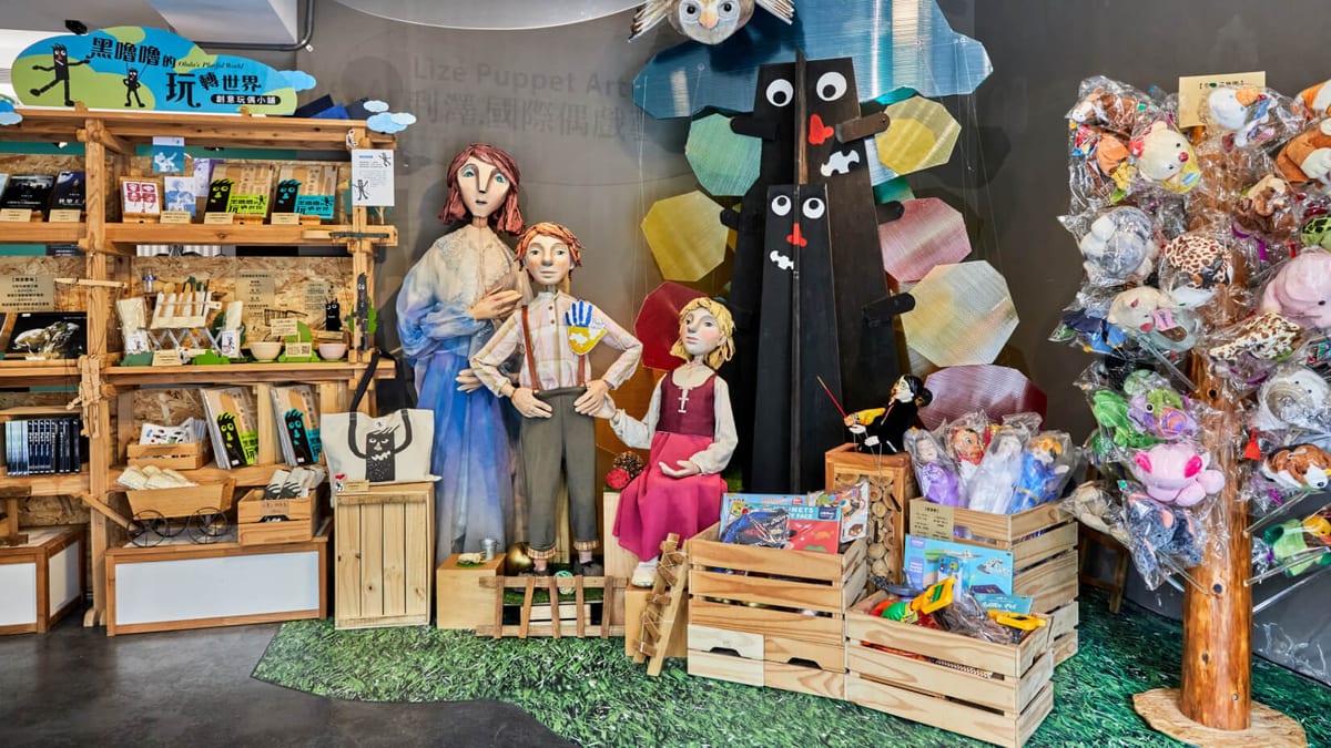 Puppets of a red-haired woman in blue, a boy with hands on his hips and a girl stand among crates and books, before a tree character with large eyes and a red nose.