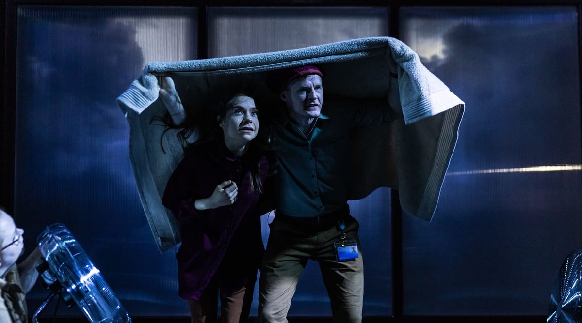 Against blue-lit translucent hospital curtains, two performers hold a grey blanket over their heads. The performer on the right wears a red cap and an identification badge.