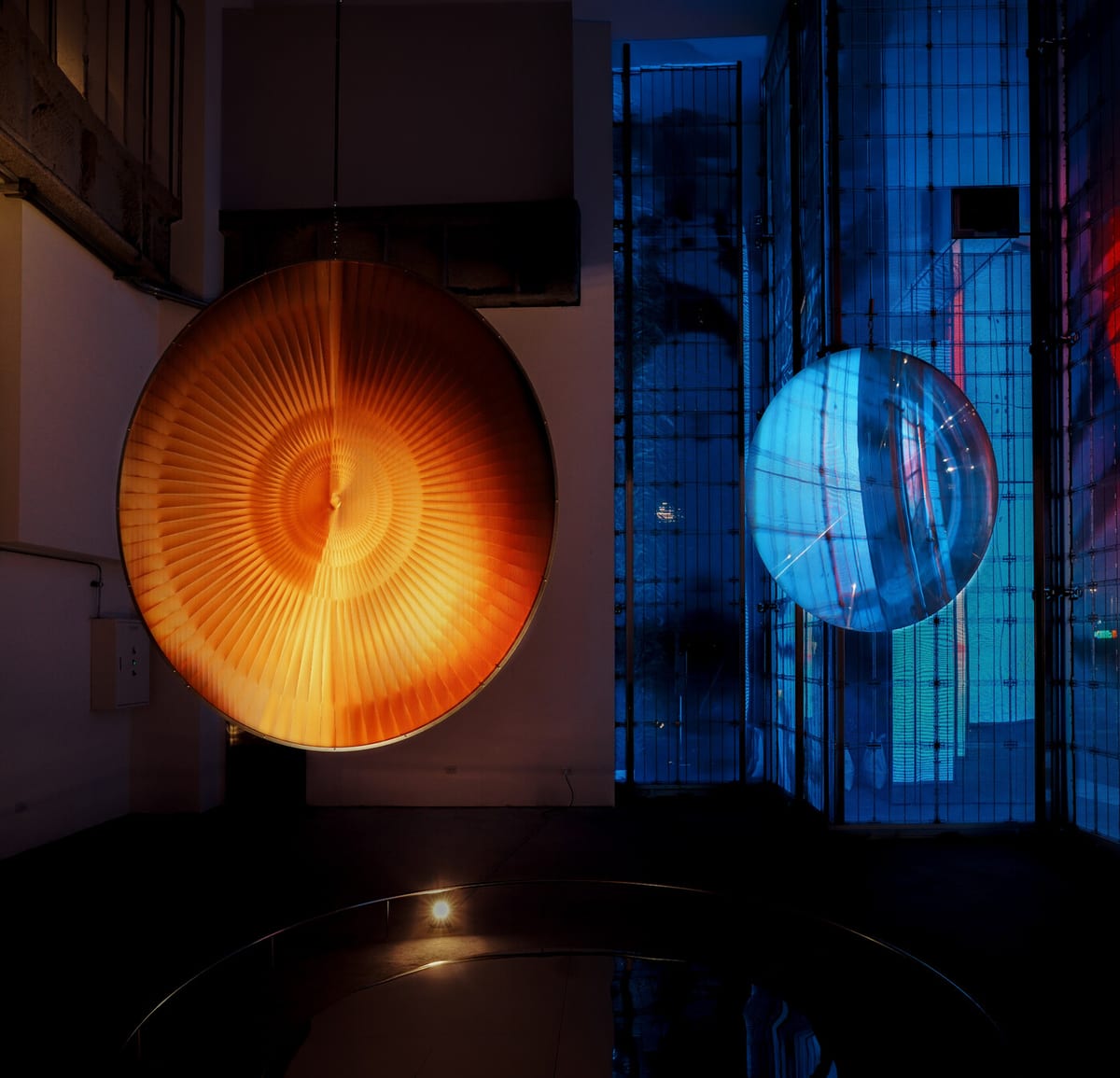 An orange disc with fine radial lines hangs beside a transparent blue-lit lens, with reflections in a dark circular mirror below.